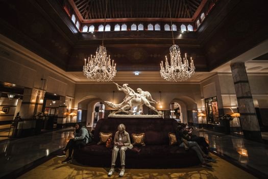 Unrecognizable people sitting on comfortable sofa in spacious hall of luxury hotel with crystal chandeliers and marble statue