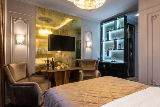 LCD television set against bed with cover in house with table between armchairs and shiny chandelier