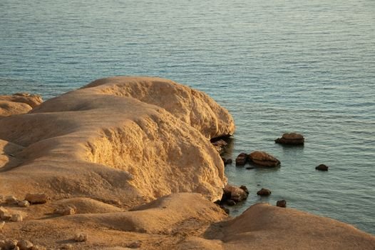 Sunlit rocky cliffs along a tranquil sea, showcasing natural erosion and serene coastal beauty.
