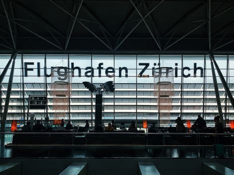 Travelers waiting at Zürich Airport with silhouettes against modern architecture.