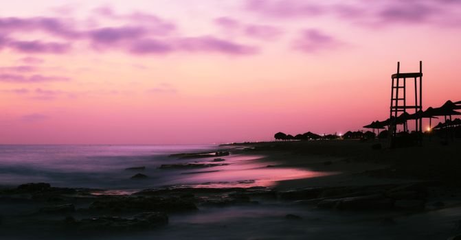 Breathtaking view of wavy ocean near stones and metal construction under picturesque colorful pink and violet clouds on sky at sundown in twilight