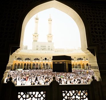 A large crowd of pilgrims surrounding the Kaaba during daylight, with a view of the Grand Mosque's architecture.
