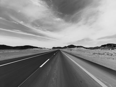 Black and white image of an empty road in Tabuk, Saudi Arabia, showcasing desert scenery and cloudy sky.